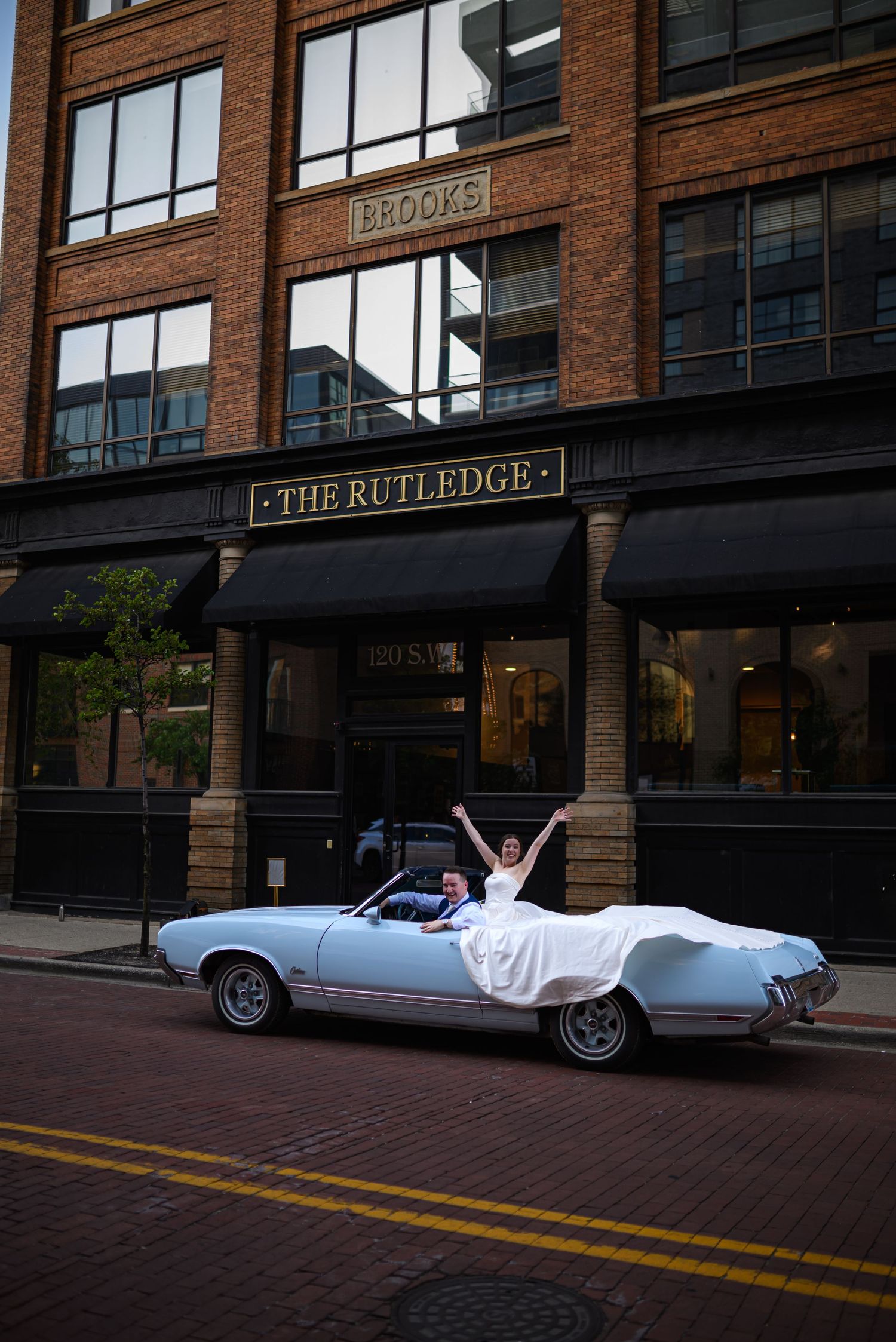 Couple poses in vintage convertible car outside brick building with storefronts.