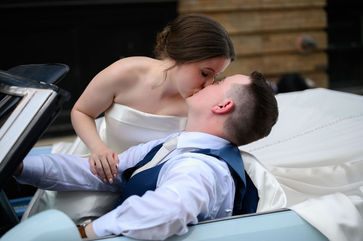 Romantic couple dressed in wedding attire share an intimate moment while reclining on a vintage car.