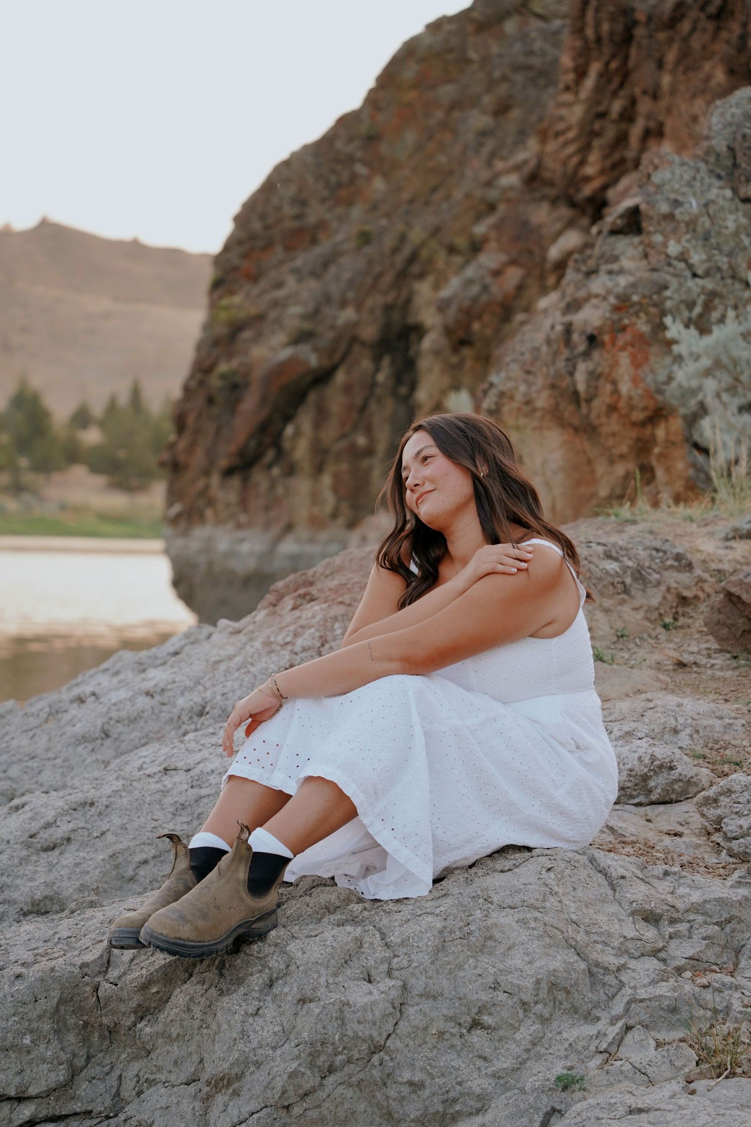 Woman in white dress and boots sitting on rocks by river at golden hour.