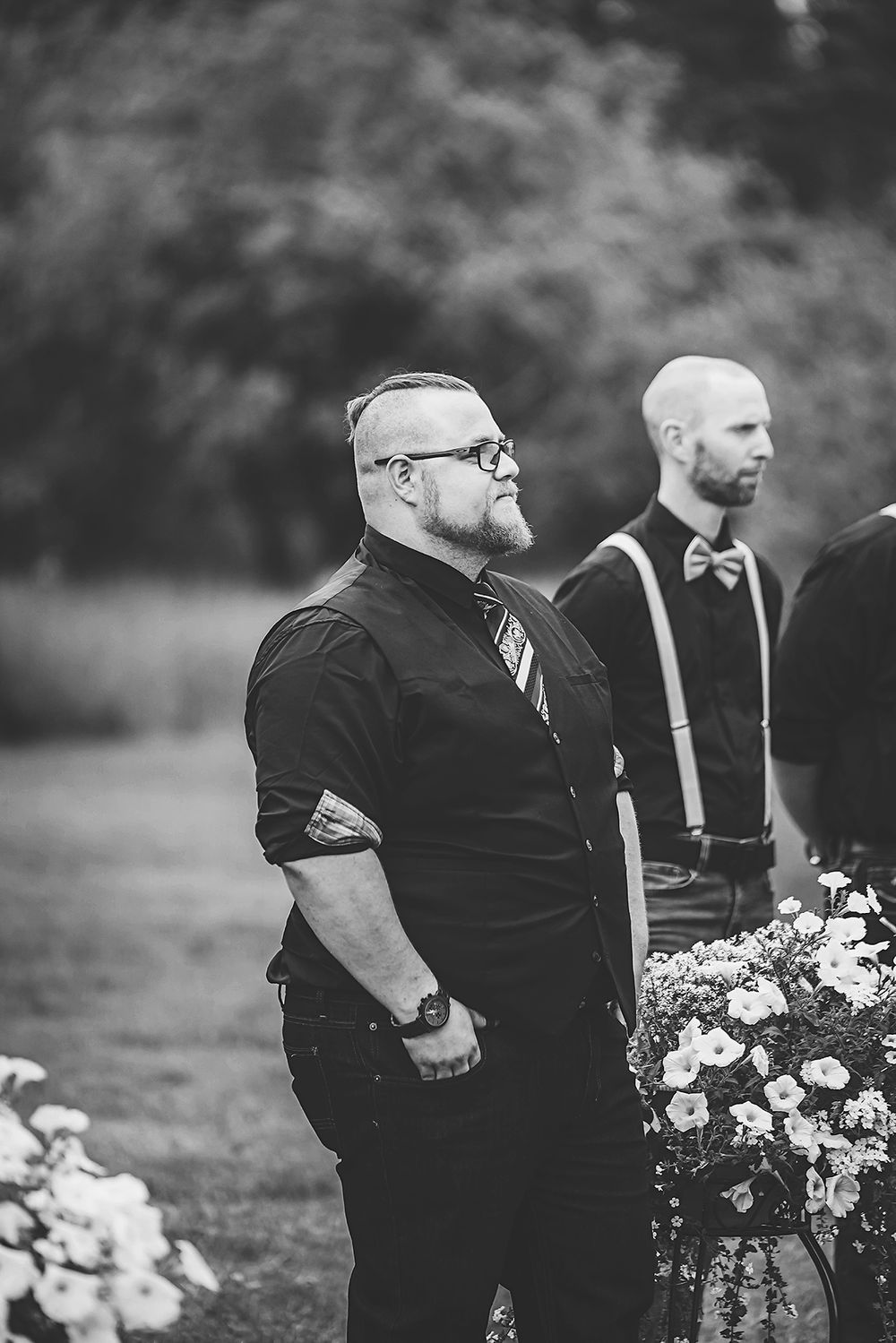 Black and white photo of groomsmen in formal attire standing beside floral arrangements at outdoor wedding ceremony.