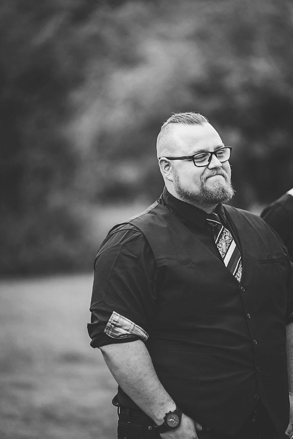 Black and white close-up portrait showing attendant in dark shirt and glasses at outdoor wedding.