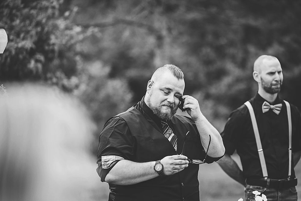 Two groomsmen in black attire and suspenders stand together during an outdoor wedding ceremony.