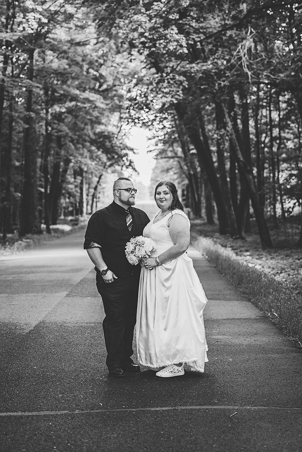 A black and white wedding portrait on a tree-lined road with natural archway.