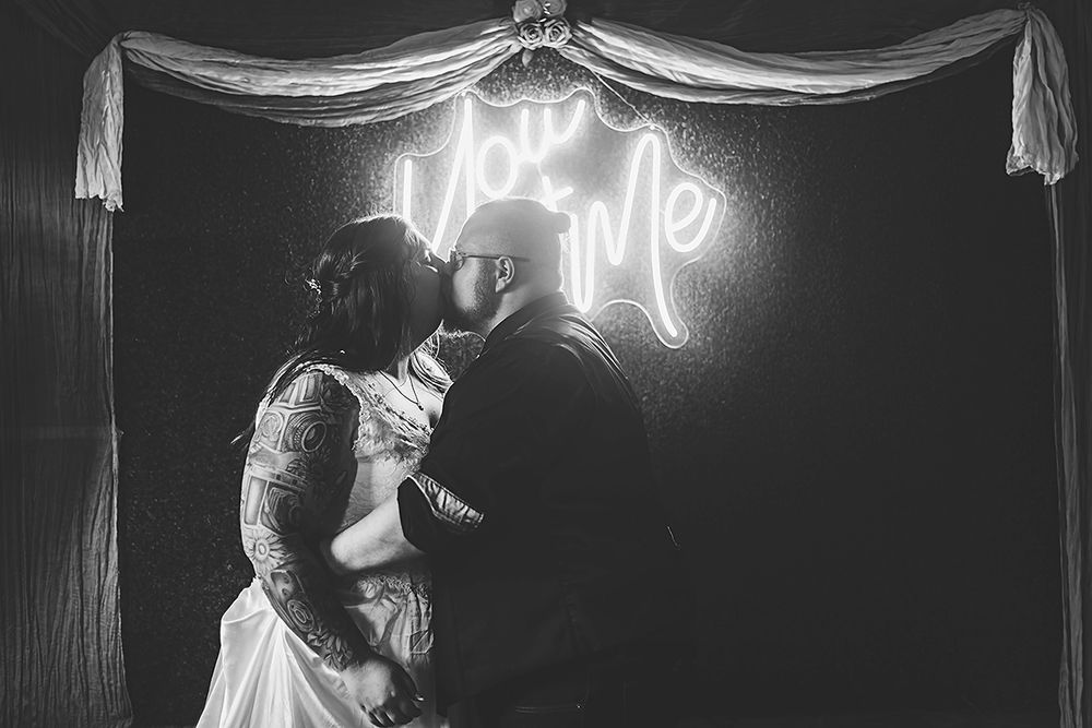 Couple sharing a romantic moment under glowing neon 'love' sign at their wedding reception in black and white.