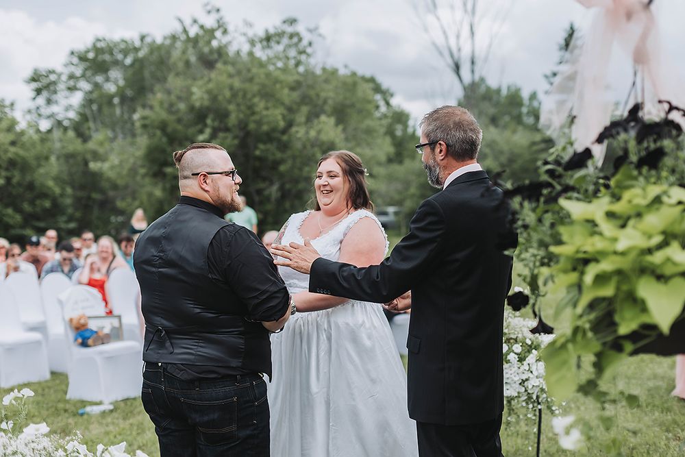 An outdoor wedding ceremony takes place in a garden setting with white chairs and greenery.