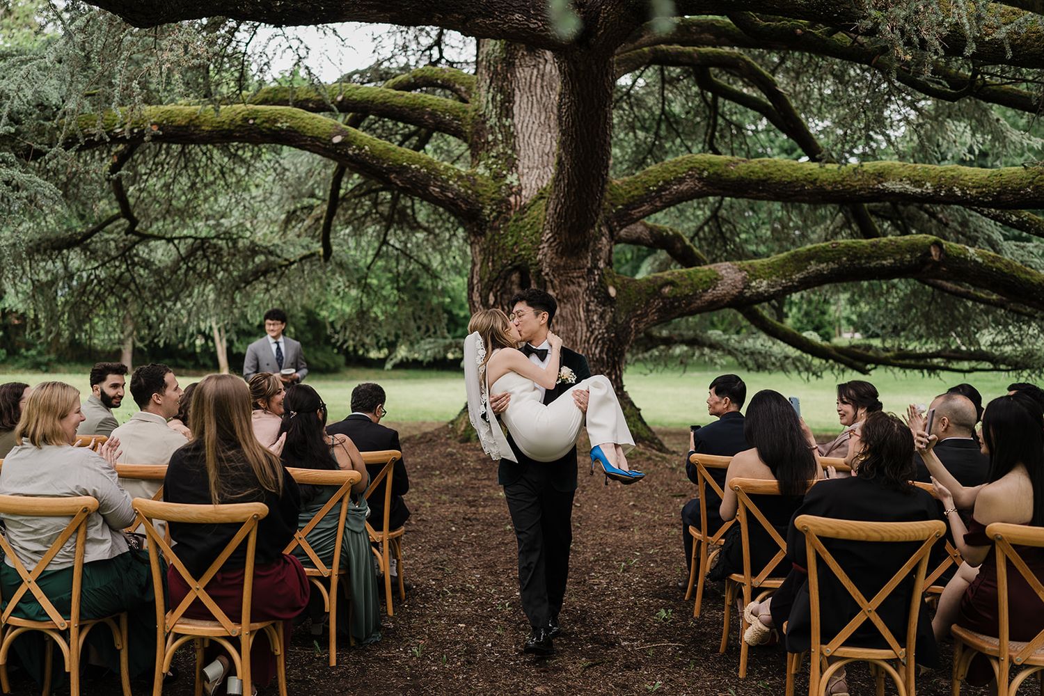 A couple embraces under a large oak tree during an outdoor wedding ceremony with wooden cross-back chairs for guests.