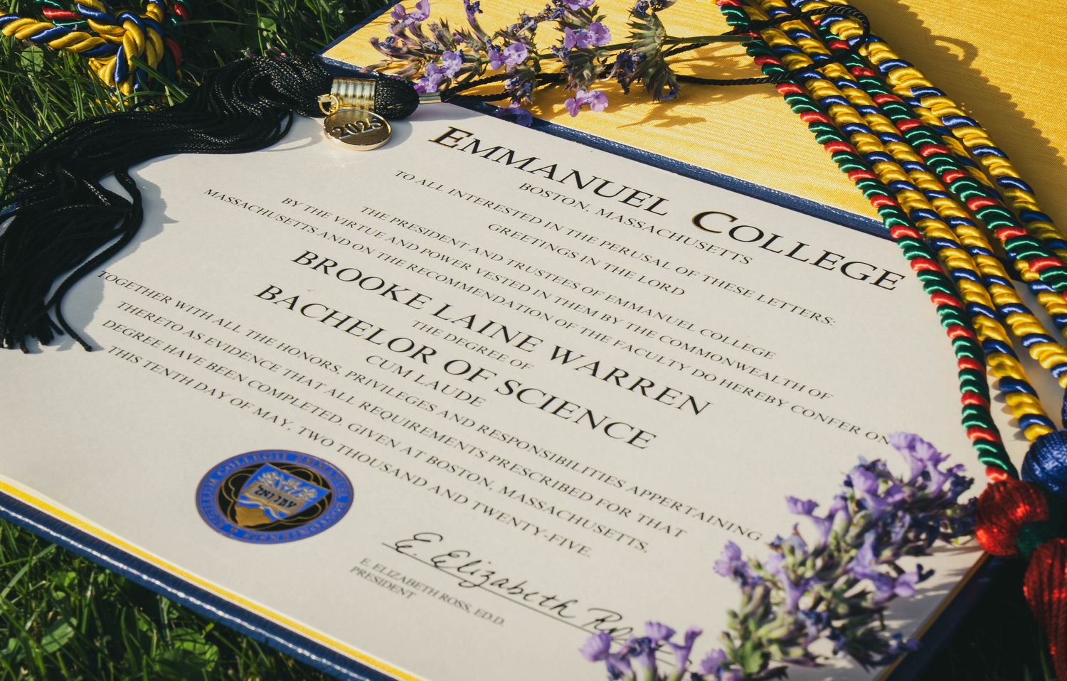 A diploma from Emmanuel College with graduation cords and decorative tassel rests on grass under golden sunlight.