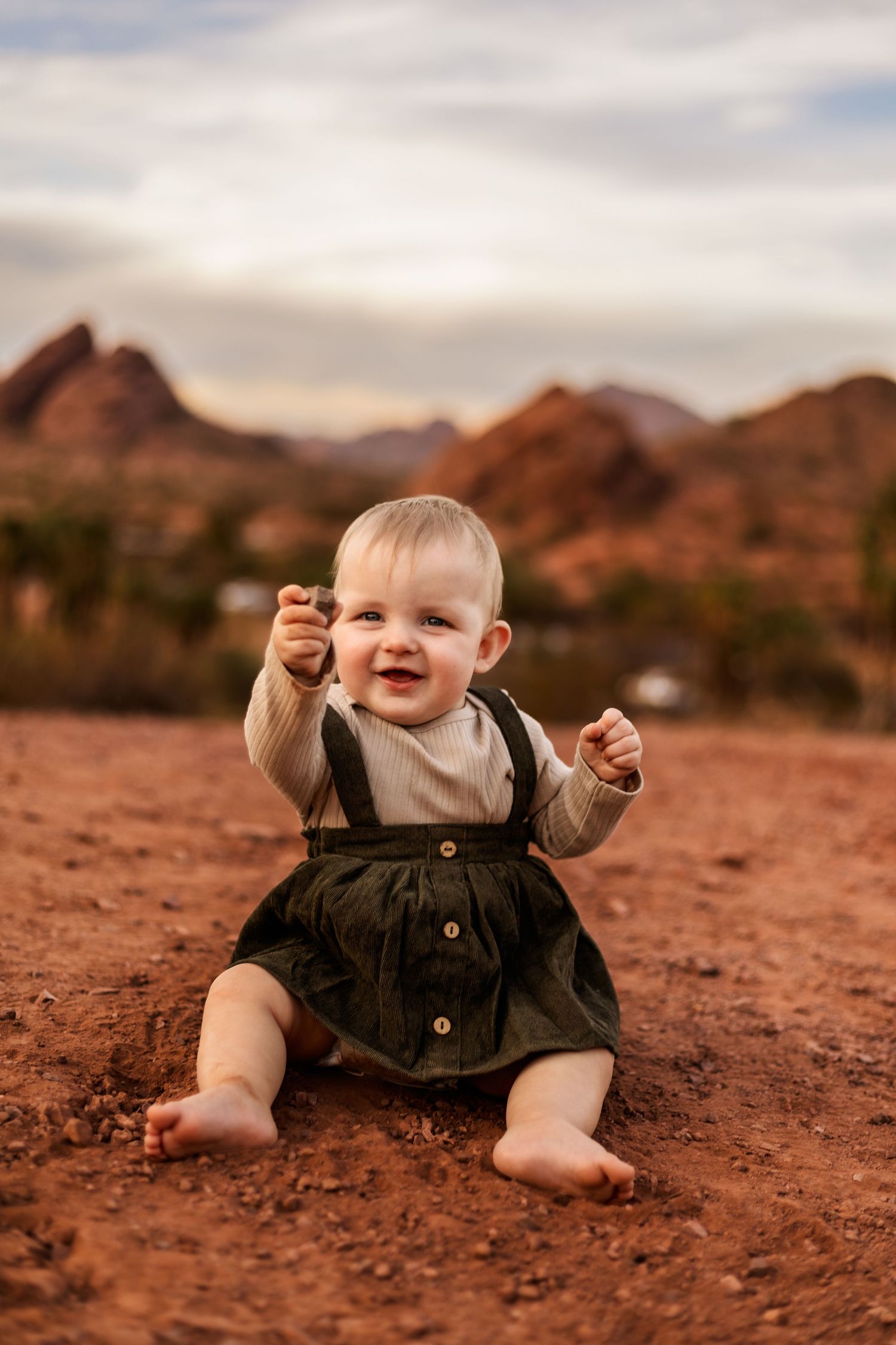 A baby wearing denim overalls and a beige shirt crawls across red desert sand against a backdrop of rocky mountains.