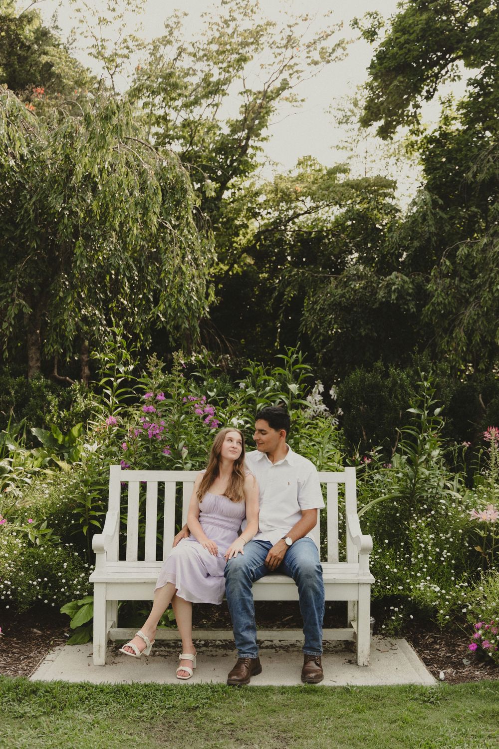 A couple sits together on a white garden bench surrounded by lush greenery and flowering plants in a peaceful setting.