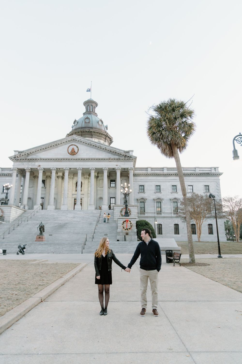 A couple holds hands in front of the grand South Carolina State House in Columbia on a cloudy day.