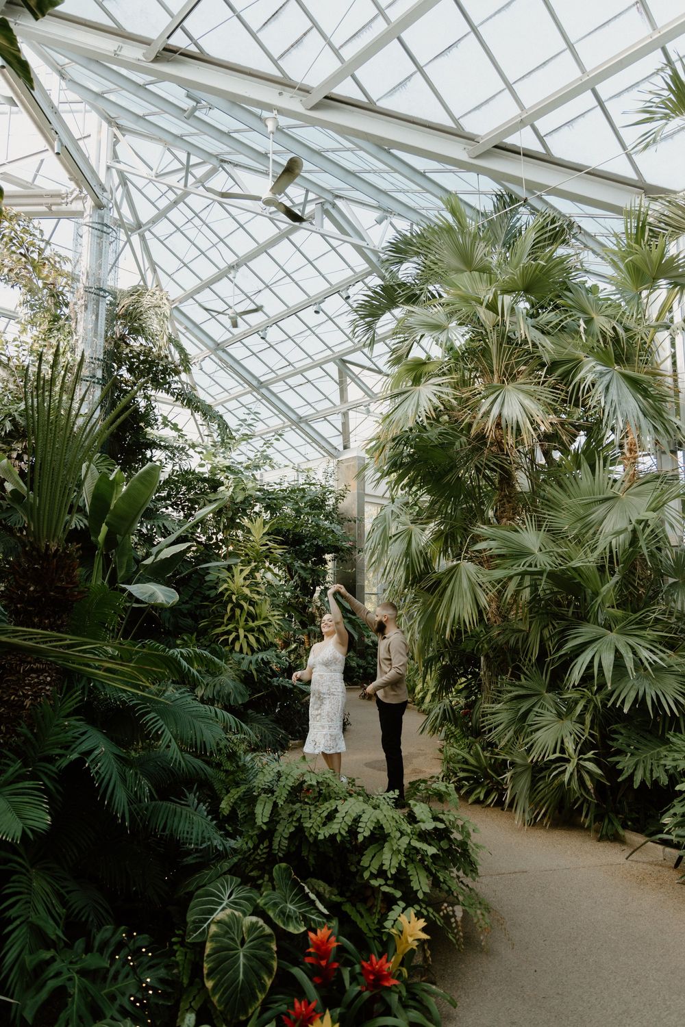 Two people stand amid lush tropical plants and palm fronds in a sunlit Victorian-style greenhouse conservatory.