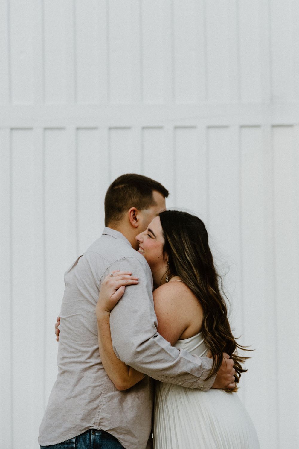 A couple embraces and kisses in front of a white textured wall while dressed in soft neutral colors.