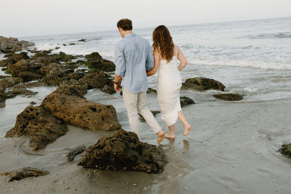 A couple in white outfits sits together on rocky beach overlooking ocean waves during golden hour.