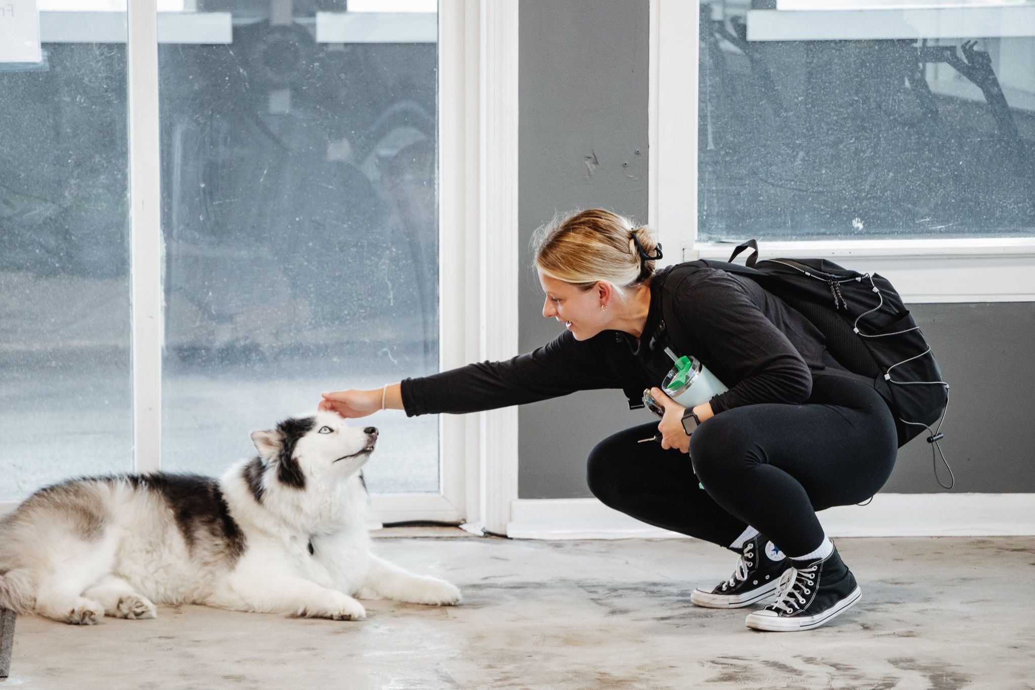 Woman crouches to gently pet a fluffy husky lying on the gym floor; she holds a shaker bottle and wears a backpack and Converse sneakers