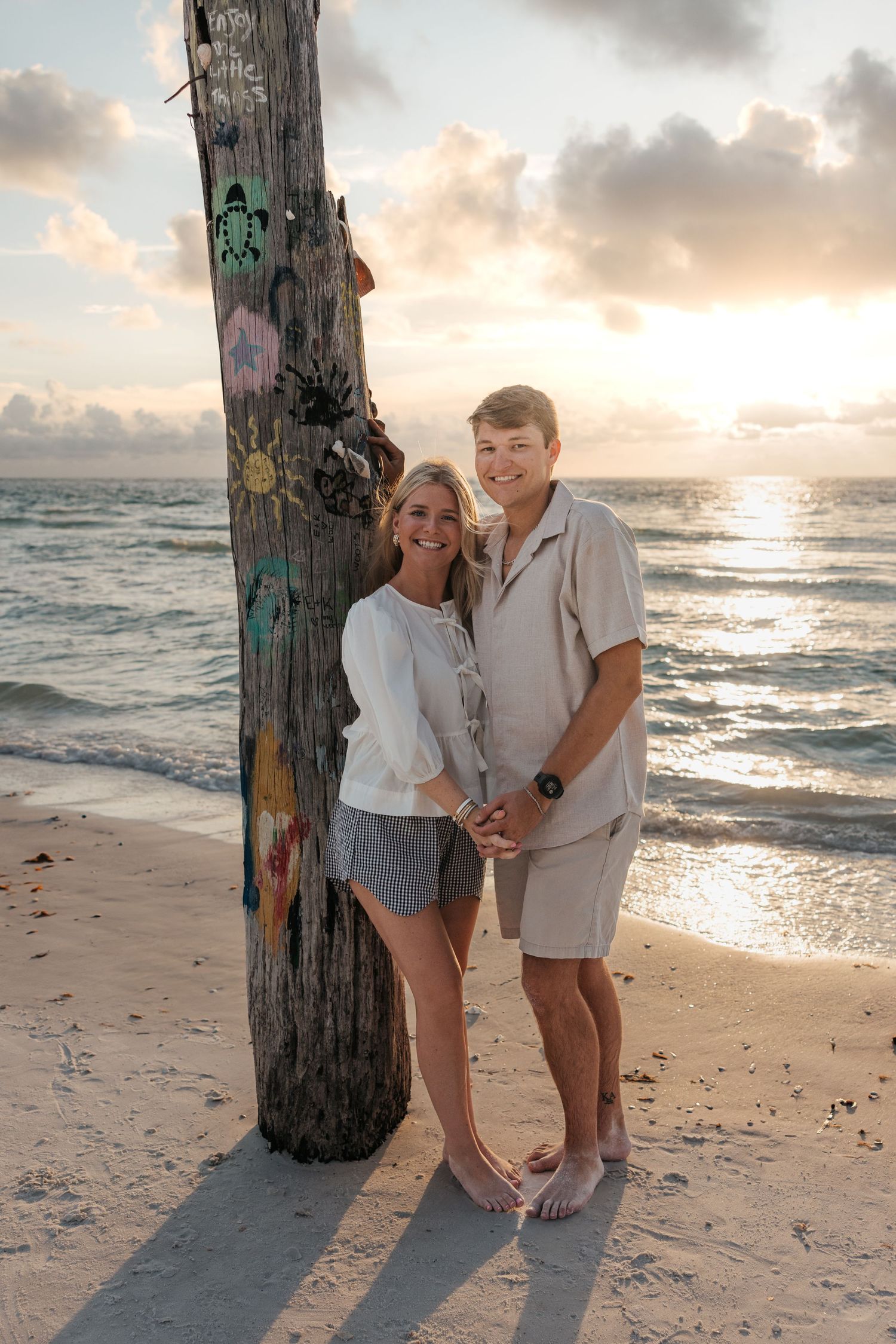 A couple poses together at sunset on a beach with graffiti covered wooden post and ocean waves in the background.