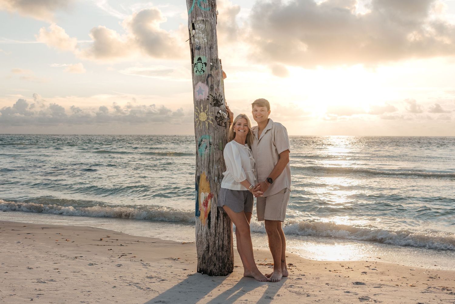 Couple embraces by palm tree on tropical beach at sunset with ocean waves and glowing sky in background.