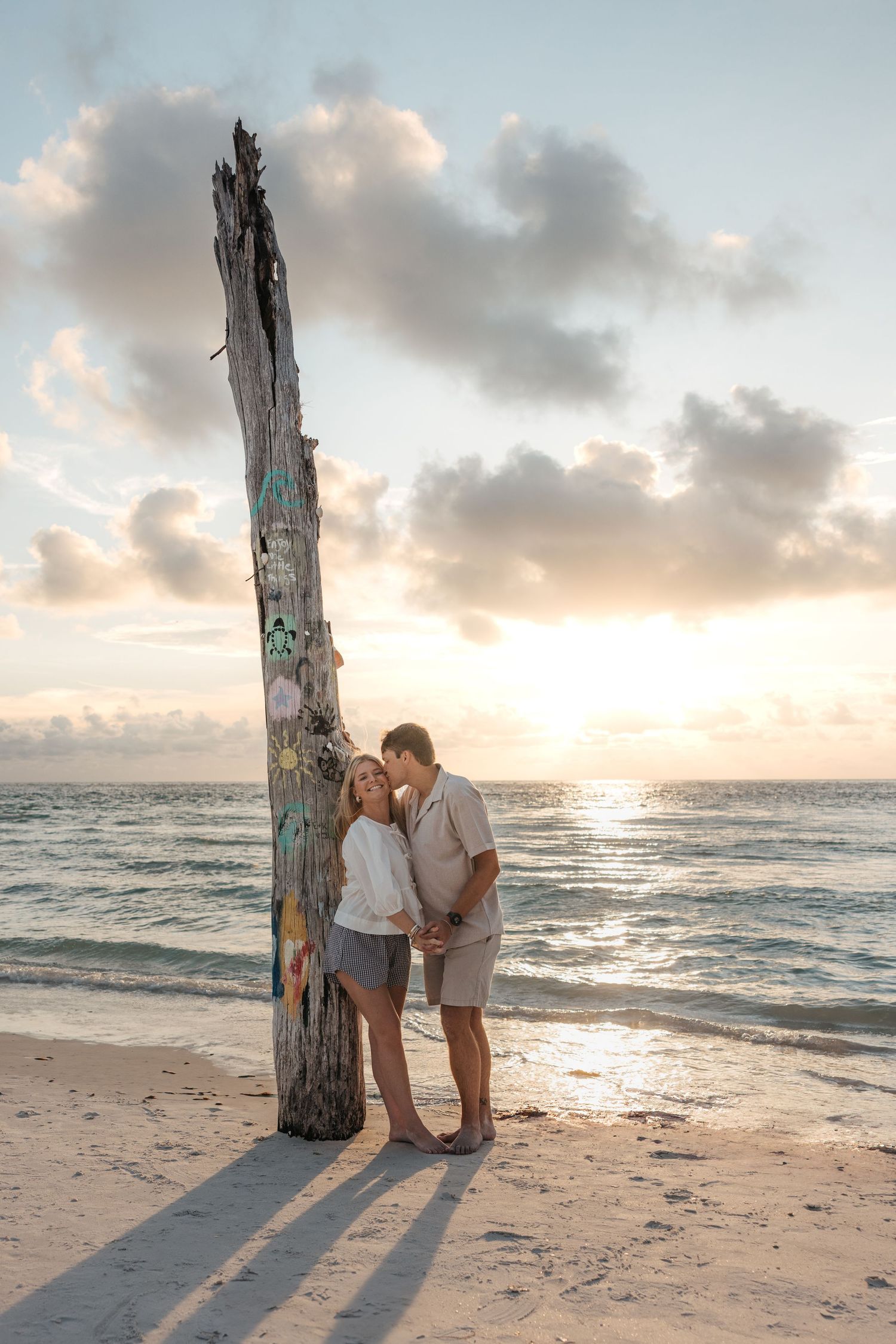 A couple shares a romantic moment at sunset on the beach next to a weathered driftwood post.