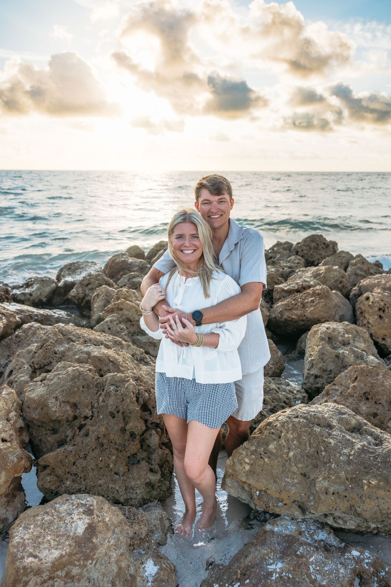 A couple shares a romantic moment on rocky coastal shore during golden hour sunset over the ocean.