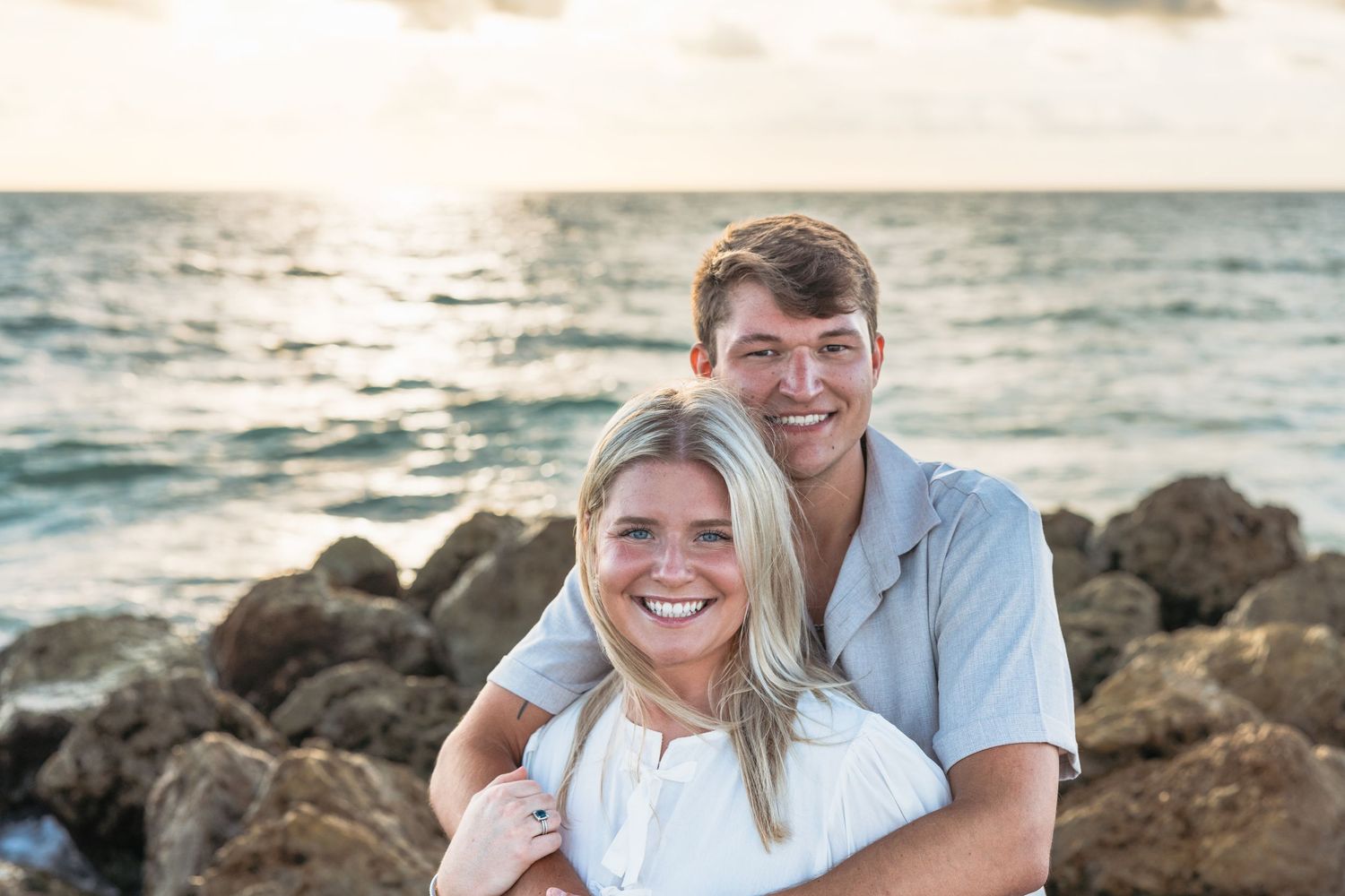 A couple smiles together on a rocky beach at sunset with ocean waves in the background.