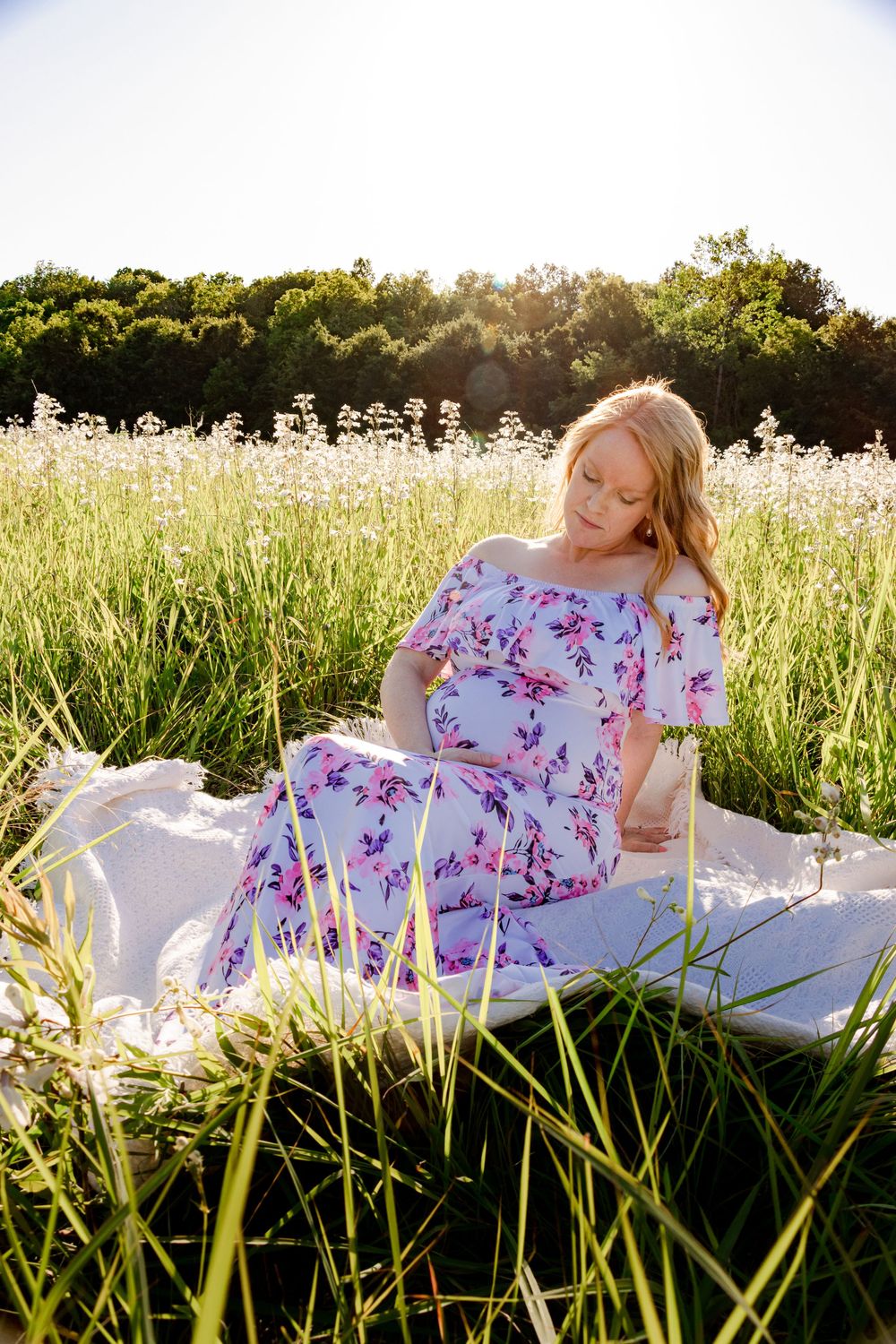 An expectant mother in a pink floral dress relaxes on a blanket in a sunny wildflower field.