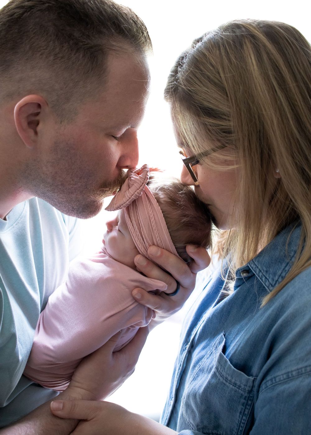 Parents share an intimate moment with their newborn baby in soft window light.