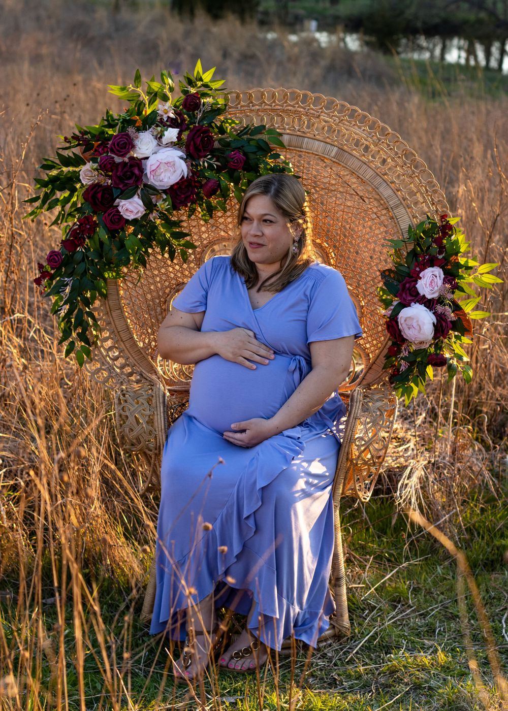 Maternity portraits in purple dress with wicker chair and floral arrangements at sunset.