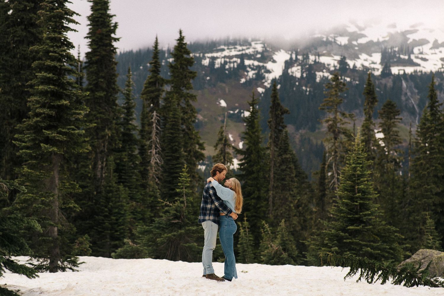 Snow-covered evergreen trees line a mountainous landscape with a couple embracing in a romantic winter scene.