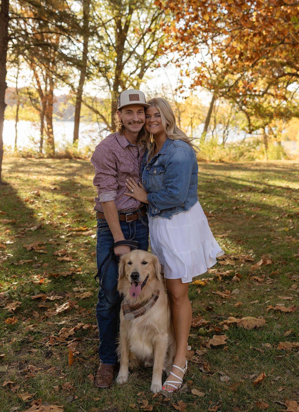 Couple poses with their golden retriever on fallen autumn leaves under trees.