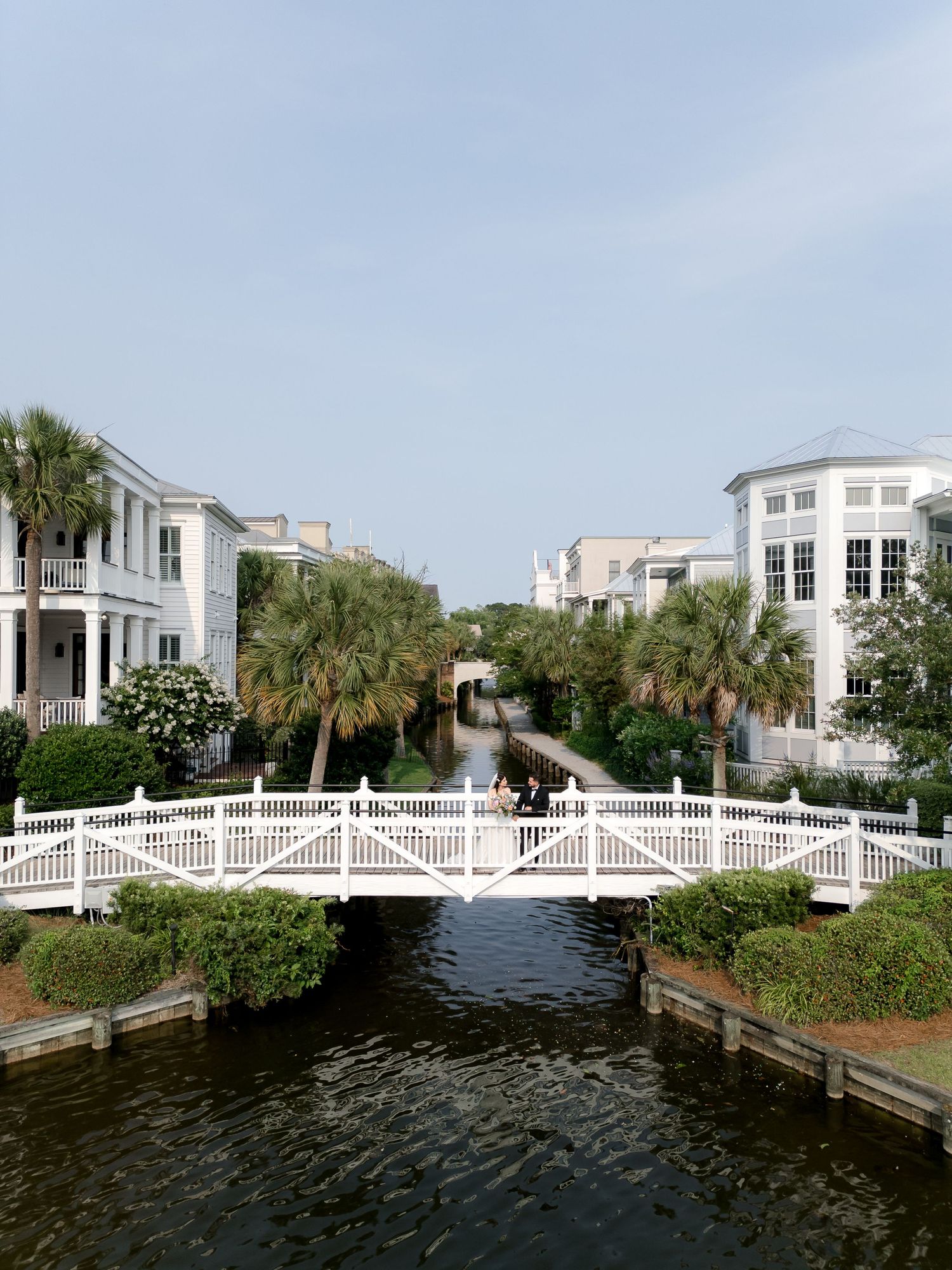 White arched bridge over a calm waterway surrounded by palm trees and elegant homes.