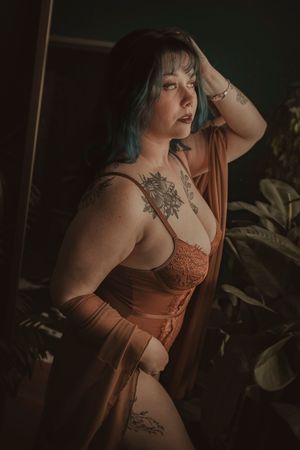 Moody portrait of person in orange lingerie with visible tattoos posing against dark background with houseplants.
