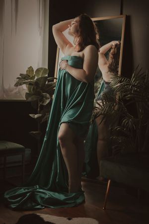 Artistic boudoir photo of woman in emerald green silk dress posing dramatically beside mirror and indoor plants.