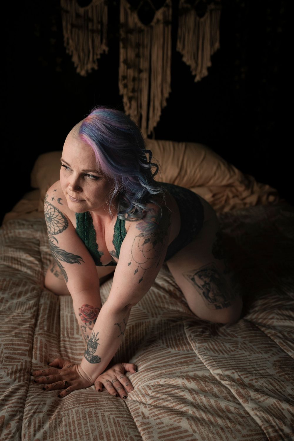 Person with blue hair and tattoos poses seductively on a quilted bed in atmospheric lighting.