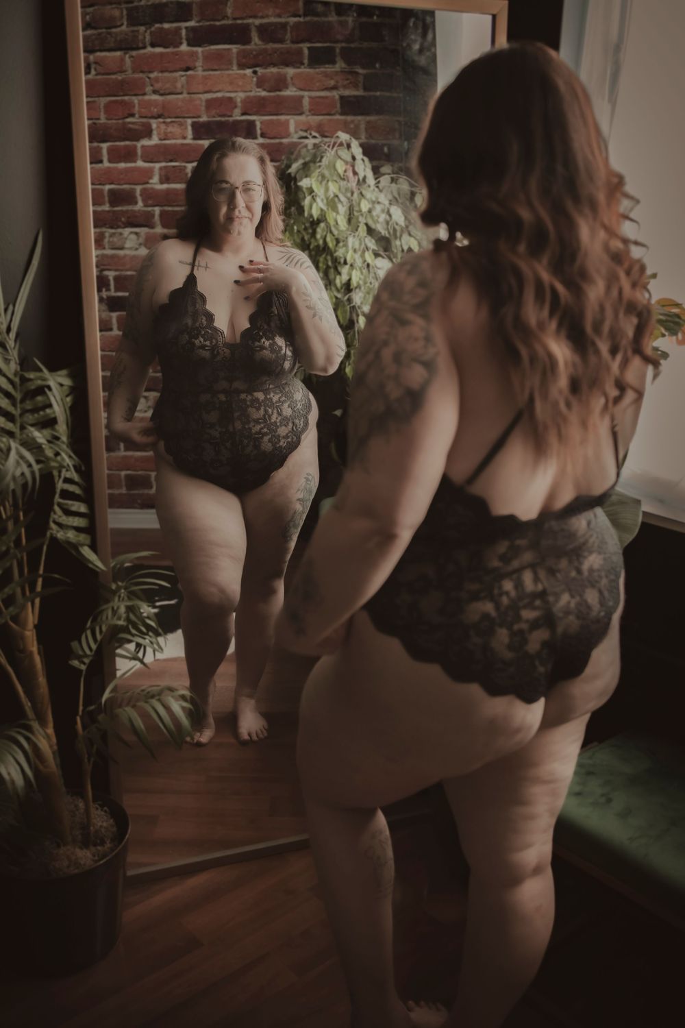 Person in black lace lingerie looking at reflection in mirror with exposed brick wall and houseplants in background.