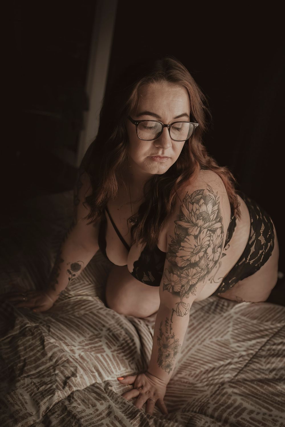 A moody boudoir portrait featuring intricate floral arm tattoos in dark tones with dramatic lighting on a patterned bed.