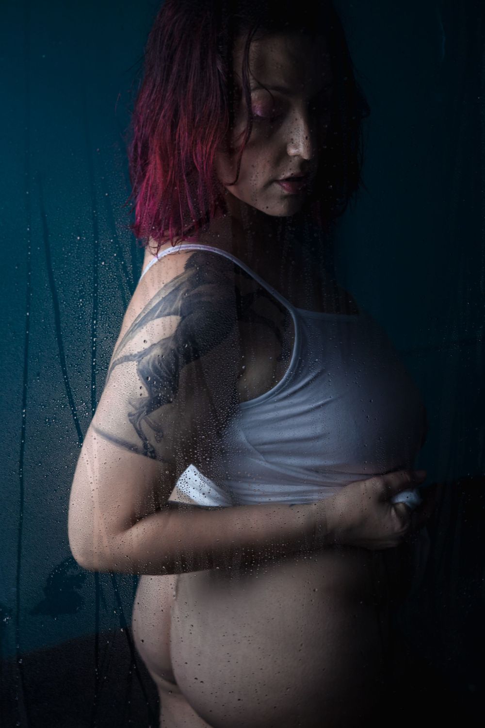 Artistic maternity photo showing a person with red hair and tattoos standing against a dark background with water droplets.