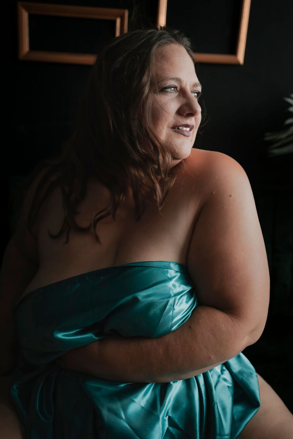 Artistic boudoir portrait in moody lighting features subject wrapped in turquoise silk fabric looking to the side.