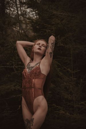 Person in rusty orange lingerie poses with arms raised in moody forest setting with visible tattoo art.