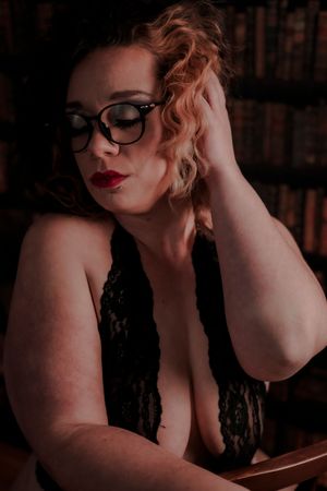 Someone in a lace top with glasses poses intimately against a bookshelf backdrop in moody lighting.