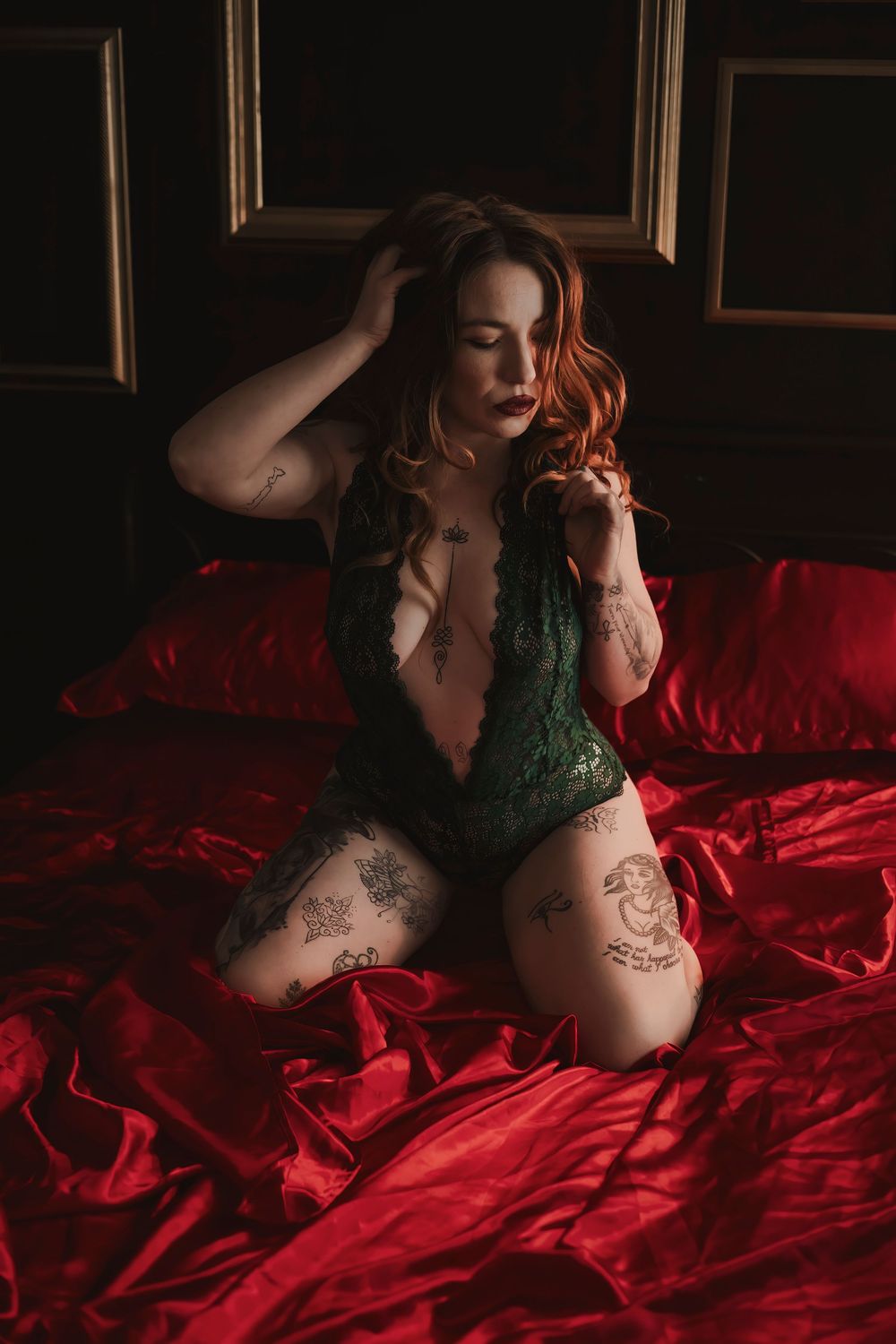 Artistic boudoir photo with green lace bodysuit against luxurious red satin bedding in moody lighting.