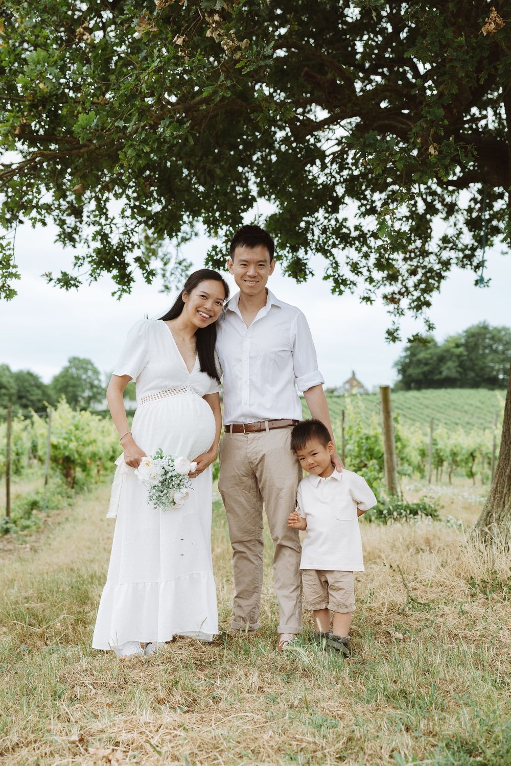 A family in white and khaki outfits poses playfully together in a sunny outdoor field with green trees in the background.