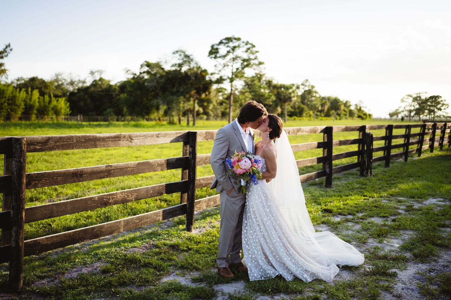 A couple shares a romantic moment at sunset by a wooden fence on a rustic farm property.