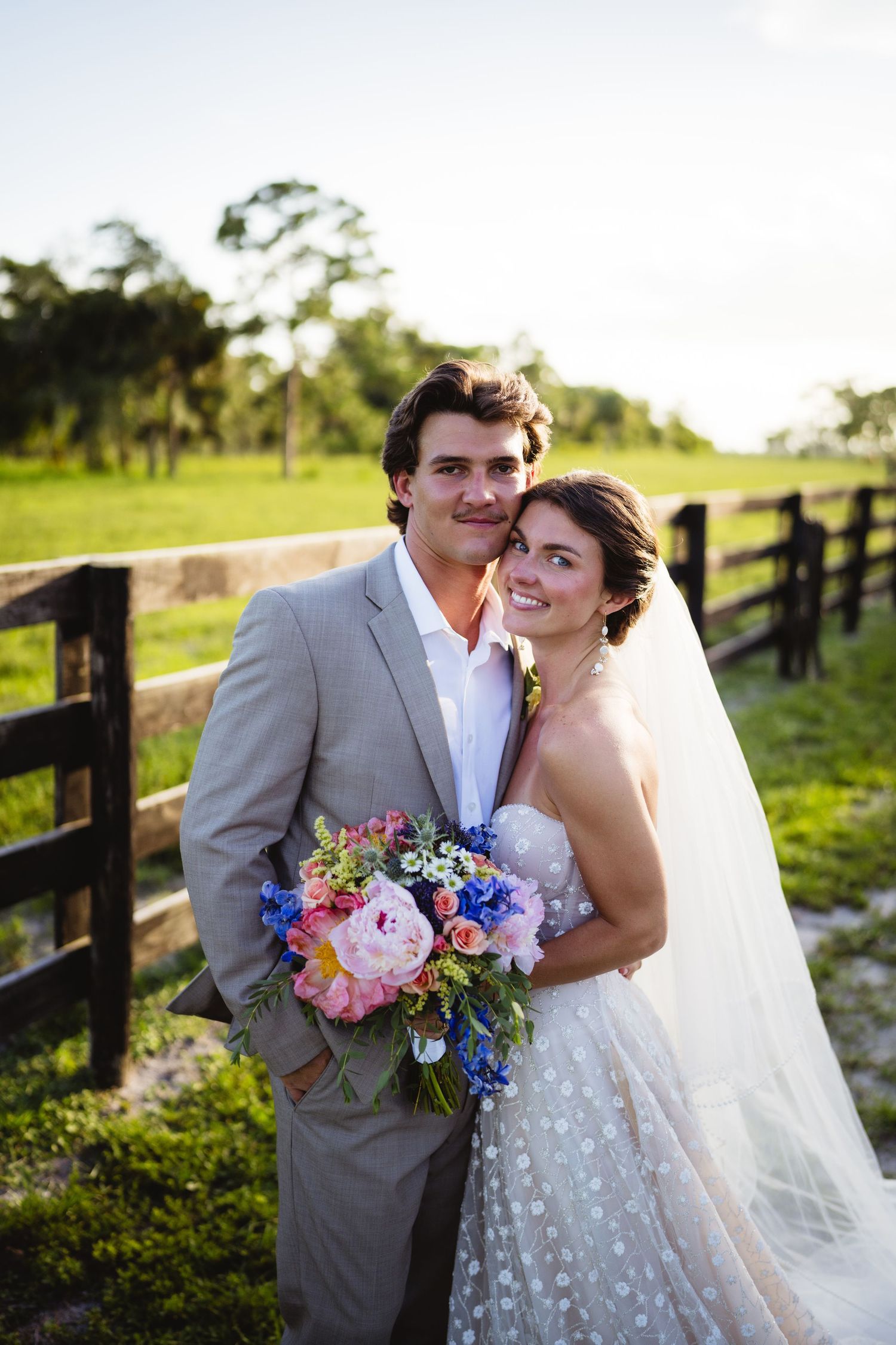 Newlyweds pose together at sunset by rustic wooden fence with colorful wedding bouquet of pink and purple flowers.