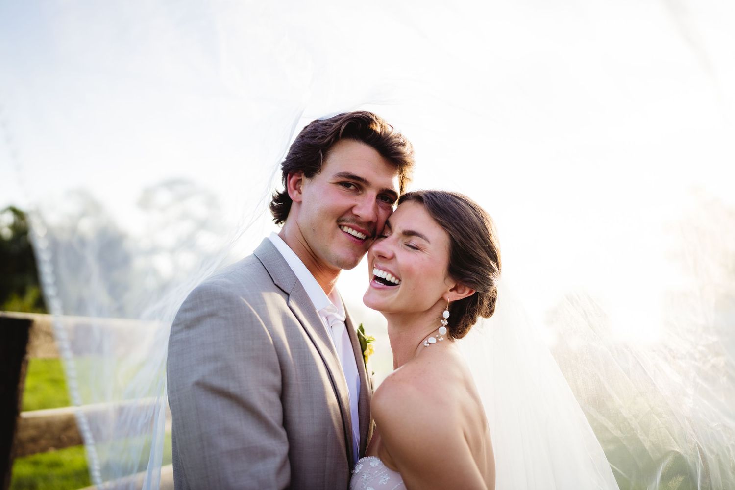 Newlywed couple shares joyful laughter together in sunlit outdoor garden setting during wedding celebration.