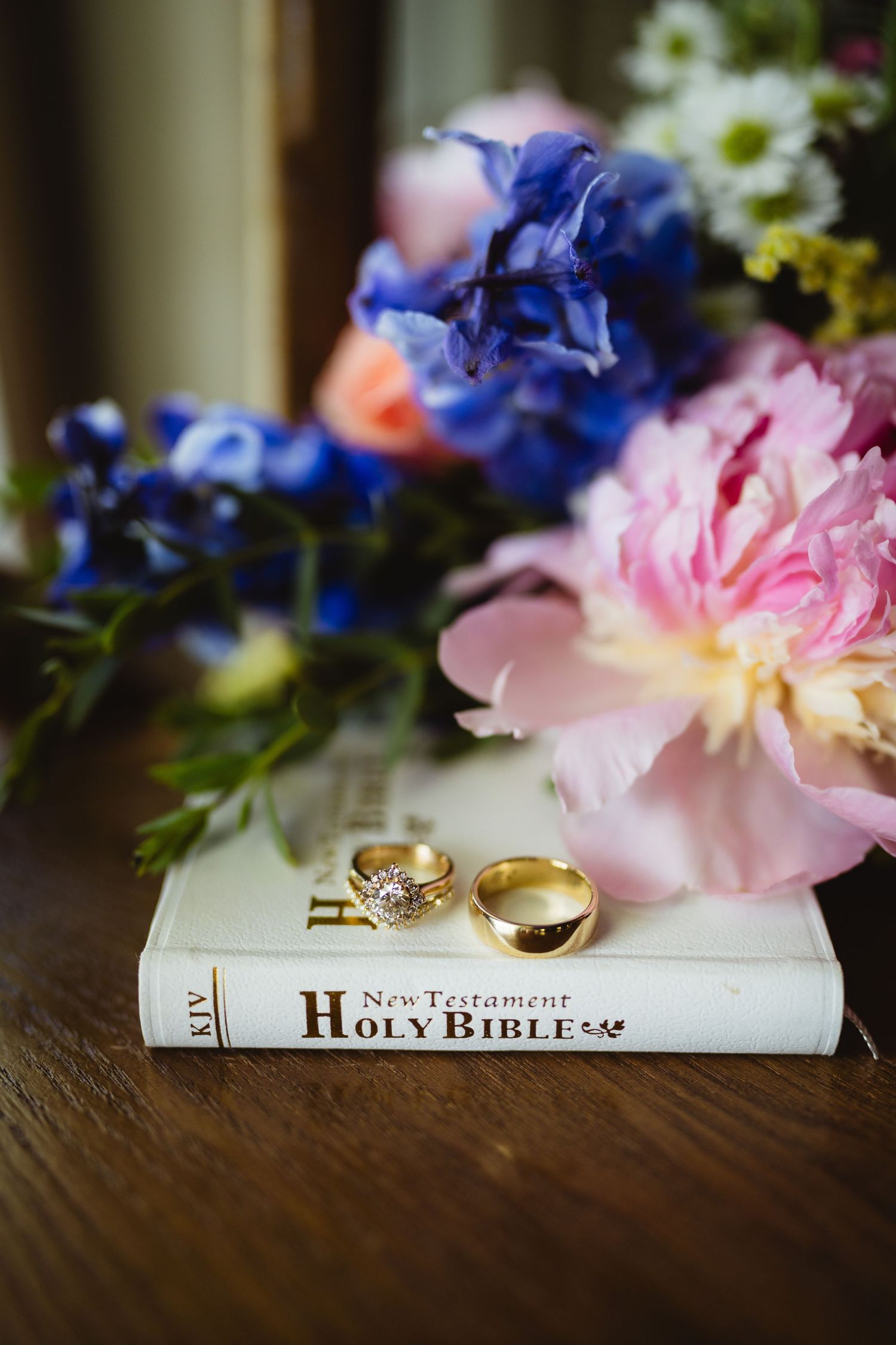 Elegant wedding ring displayed on driftwood amid vibrant blue delphiniums, pink roses and greenery in a floral arrangement.