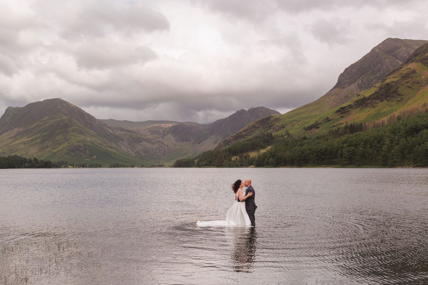 A dramatic wedding photo of a couple standing in a lake with mountains and moody skies behind them.