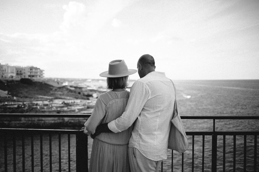 A couple in summer attire stands together looking out over a railing at a scenic coastal landscape in black and white.