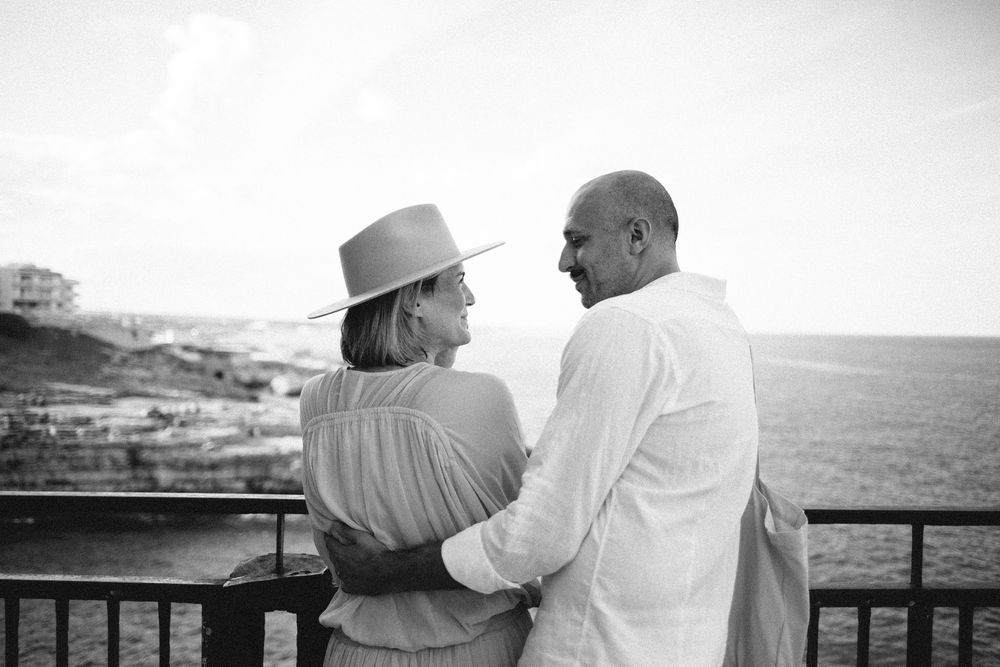 A couple in casual attire stands together on a balcony overlooking a scenic coastal landscape in black and white.