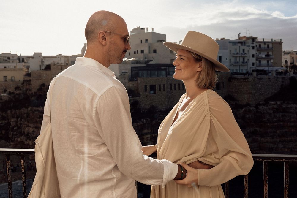 Two people in light colored clothing share an intimate moment on a balcony overlooking ancient stone buildings in a desert city.