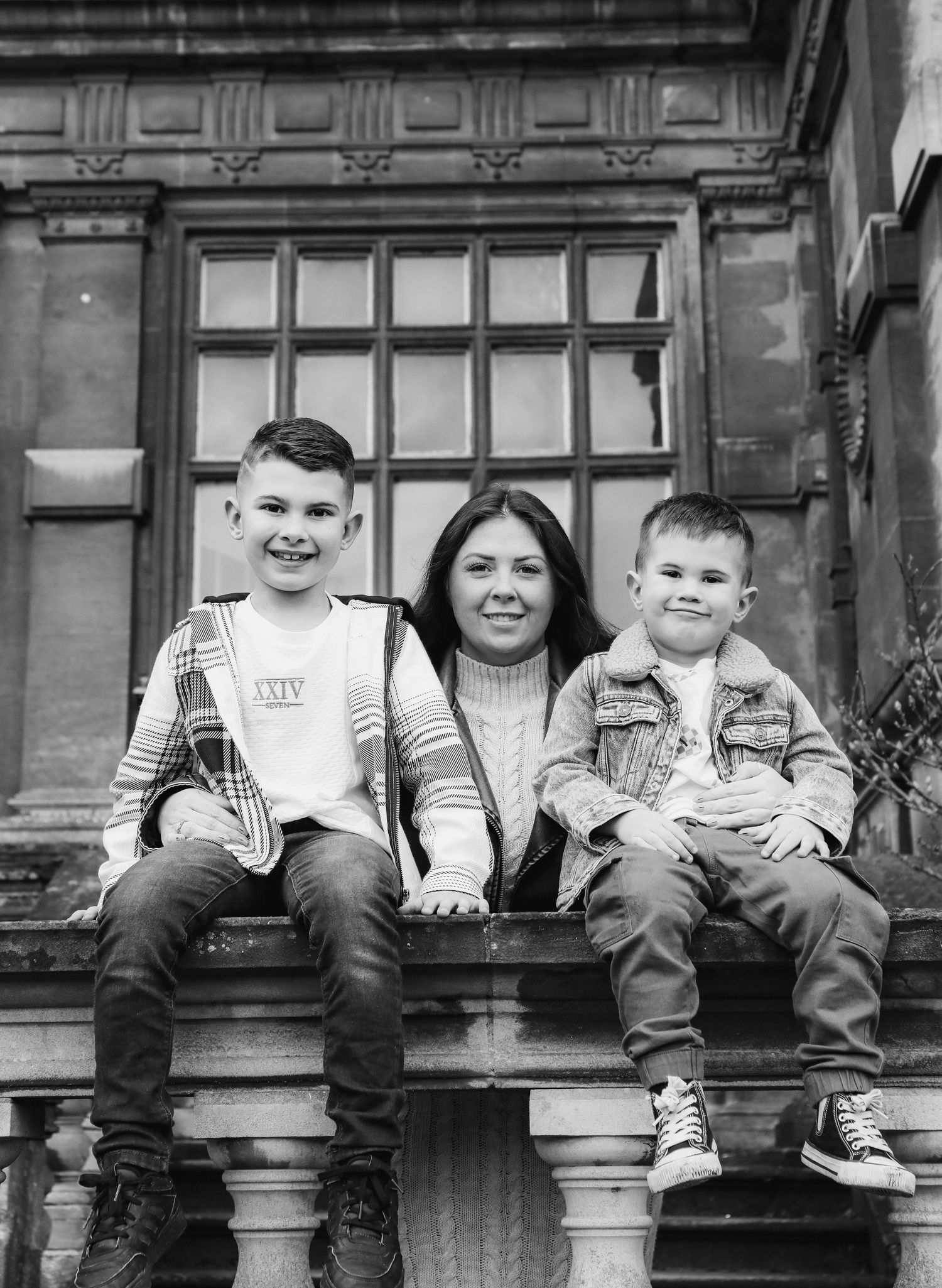 Family sat together smiling on a decorative stone ledge outside a building with large windowpanes, during family shoot in Nottingham