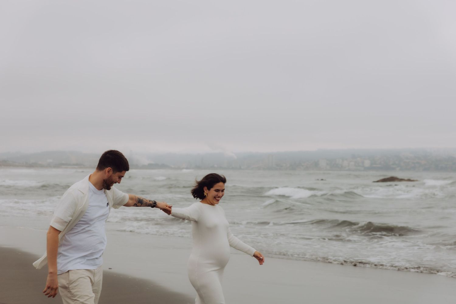 Pregnant couple in white outfits walks hand-in-hand along a stormy ocean beach.